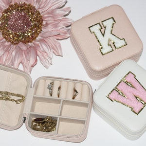 Peut inclure: Deux petites boîtes à bijoux roses et blanches avec des lettres en paillettes dorées. Les boîtes sont carrées et ont un couvercle à charnière. La boîte rose a la lettre "K" sur le couvercle et la boîte blanche a la lettre "W" sur le couvercle. Les boîtes sont ouvertes et montrent les compartiments intérieurs pour ranger les bijoux.