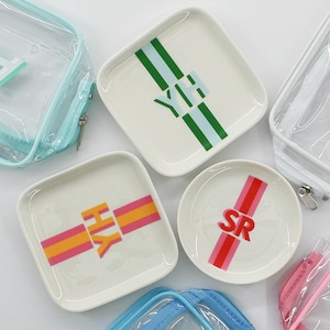 May include: Set of five clear plastic zippered pouches with white trim and colorful personalized trays. The trays have white backgrounds with colorful stripes and initials in the center. The initials are "AH", "YH", "HY", "SR", and "SE".