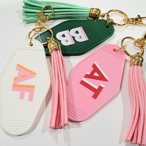 May include: Three keychains with pink, green and white motel key-shaped tags. Each tag has a different letter combination: "AF", "BB", and "TA". The keychains have pink and green tassels.