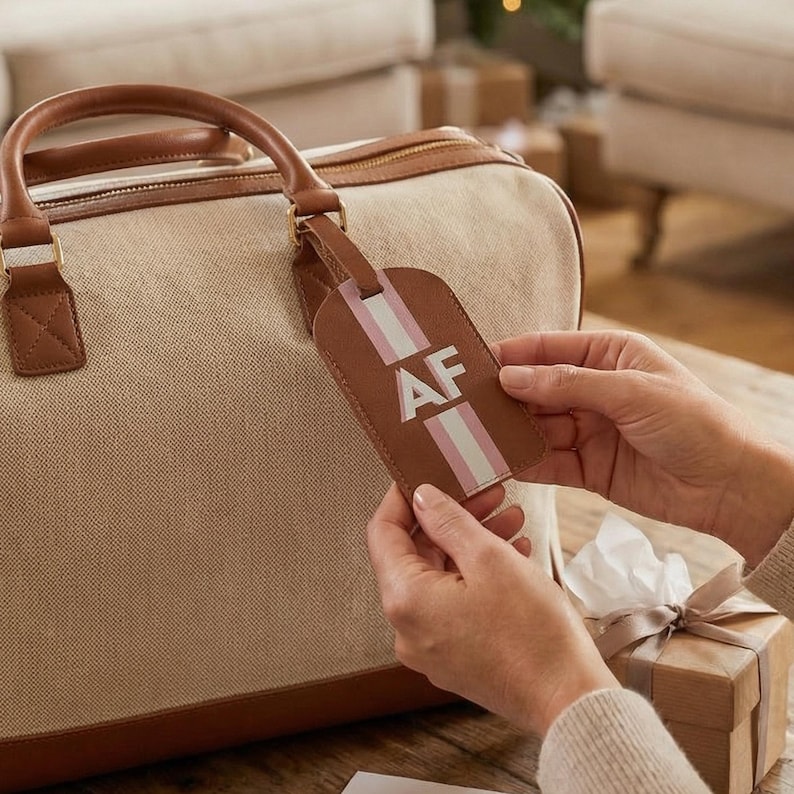May include: A tan canvas duffel bag with brown leather handles and trim, featuring a matching brown leather luggage tag with a vertical pink and white stripe and the white capital letters AF embossed on the center of the tag.