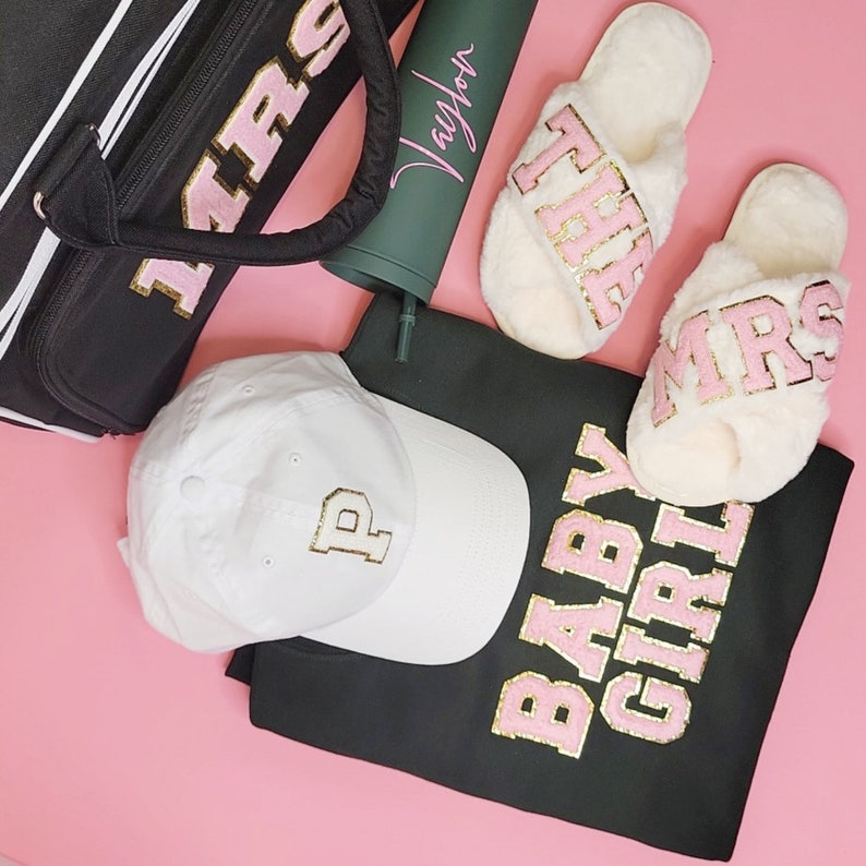 May include: A collection of accessories, including a black tote bag, a white baseball cap with a gold "P", a green tumbler with "Taylor" in pink, fluffy slippers with "THE MRS" in pink, and a black sweatshirt with "BABY GIRL" in pink.
