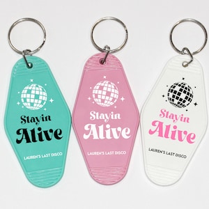 May include: Three keychains with a retro motel key design. Each keychain features a white disco ball graphic and the text "Stayin Alive" in black and pink lettering. The keychains are turquoise, pink, and white.