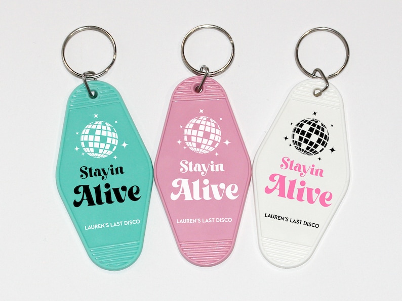 May include: Three keychains with a retro motel key design. Each keychain features a white disco ball graphic and the text "Stayin Alive" in black and pink lettering. The keychains are turquoise, pink, and white.