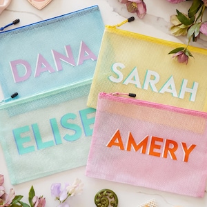 May include: Four colorful mesh zipper pouches, each with a name in bold, block letters: DANA, ELISE, SARAH, and AMERY. The pouches are in shades of blue, yellow, and pink, with black zippers. The background includes flowers and makeup.