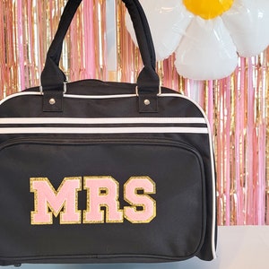 May include: Black duffel bag with white stripes and handles. The word "MRS" is displayed in pink and gold lettering on the front. The bag has a zippered front pocket and a white trim.