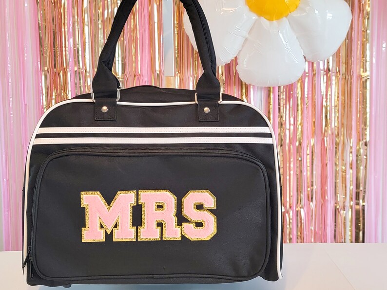May include: Black duffel bag with white stripes and handles. The word "MRS" is displayed in pink and gold lettering on the front. The bag has a zippered front pocket and a white trim.