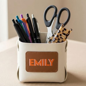 May include: A cream-colored desk organizer with the name "EMILY" in orange and pink lettering on a brown patch. The organizer holds pens, scissors, and a tortoiseshell hair clip.