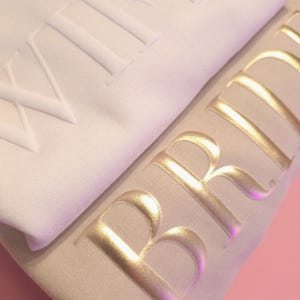 May include: Two white sweatshirts with the words "Wife" and "Bride" in raised lettering. The "Bride" sweatshirt has gold lettering.