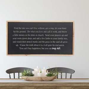 May include: A black framed sign with a white background and a quote about finding love, working hard, and growing a family. The sign is hanging on a white wall above a wooden table with four gray chairs. The table is set with a wooden bowl filled with green plants and white candles.