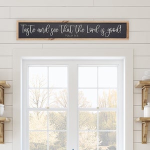 May include: A bright, inviting kitchen scene with white cabinetry, wooden shelves, and a farmhouse sink. A sign above the window reads "Taste and see that the Lord is good!" The shelves display white dishes, bowls, and decorative items. Two black sconces flank the window.