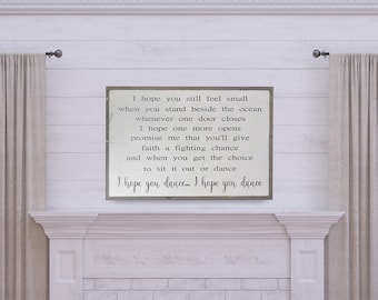 May include: A white wooden sign with a gray background and black text. The text reads: "I hope you still feel small when you stand beside the ocean whenever one door closes I hope one more opens promise me that you'll give faith a fighting chance and when you get the choice to sit it out or dance I hope you dance. I hope you dance."