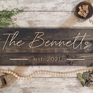 May include: A dark brown wooden sign with the text "The Bennetts est. 2021" in gold lettering. The sign is made of reclaimed wood and has a rustic look.