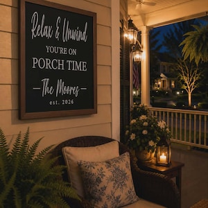 May include: A cozy porch scene at dusk. A framed sign reads "Relax & Unwind, You're on Porch Time - The Moores, est. 2026." A wicker chair with patterned pillows sits near a small table with a lit candle and lantern. Lanterns hang from the ceiling.