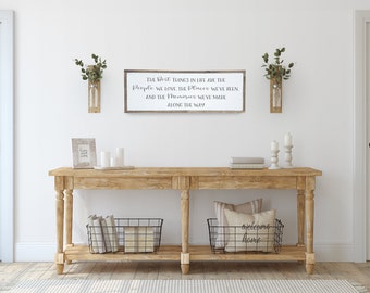 May include: A wooden sign with the text "The best things in life are the people we love, the places we've been, and the memories we've made along the way." The sign is hanging on a white wall above a wooden console table with two black wire baskets, one with the text "welcoming home".