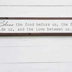 May include: A wooden sign with a brown frame and white background. The sign reads "Bless the food before us, the family beside us, and the love between us. Amen."