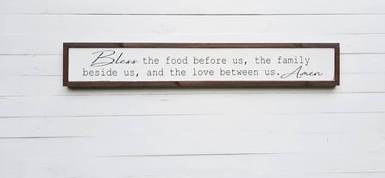 May include: A wooden sign with a brown frame and white background. The sign reads "Bless the food before us, the family beside us, and the love between us. Amen."