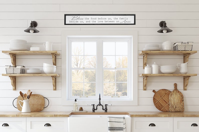 May include: A white kitchen with wooden shelves, a window, and a farmhouse sink. A black framed sign above the window reads "Bless the food before us, the family beside us, and the love between us. Amen."