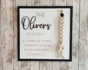 May include: A framed white sign with black lettering and a macrame accent. The sign reads "THE Olivers" above the date "05.23.2023" and a quote from Ecclesiastes 4:12. A beige macrame cord is attached to the right side.