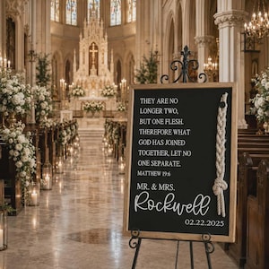 May include: A rectangular black wedding sign in a light wood frame on a black metal easel. The sign features white text reading: THEY ARE NO LONGER TWO, BUT ONE FLESH. THEREFORE WHAT GOD HAS JOINED TOGETHER, LET NO ONE SEPARATE. MATTHEW 19:6 MR. & MRS. Rockwell 02.22.2025. A thick white rope is attached to the right side.