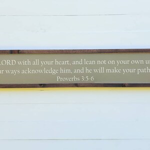 May include: A wooden sign with a beige background and a brown frame. The sign features a white inscription with the text "Trust in the LORD with all your heart, and lean not on your own understanding; in all your ways acknowledge him, and he will make your path straight. Proverbs 3:5-6".