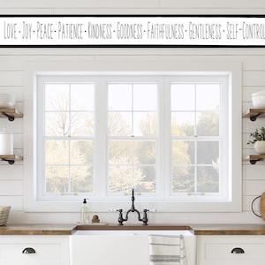 May include: A white kitchen with a window and two shelves on each side. The shelves are filled with dishes, plants, and other kitchen items. Above the window is a black sign with the text "LOVE - JOY - PEACE - PATIENCE - KINDNESS - GOODNESS - FAITHFULNESS - GENTLENESS - SELF-CONTROL".