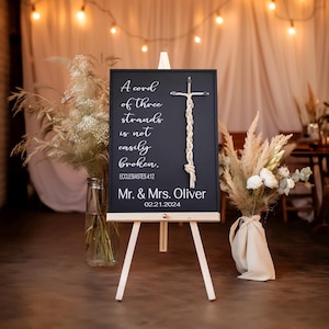 May include: A black chalkboard sign on a wooden easel, featuring white text and a macrame cross. The sign reads "A cord of three strands is not easily broken. Mr. & Mrs. Oliver 02.21.2024". Decorative floral arrangements flank the sign.
