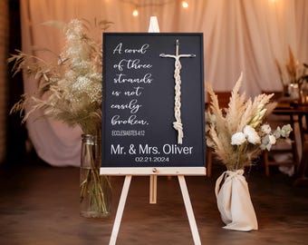 May include: A black chalkboard sign on a wooden easel, featuring white text and a macrame cross. The sign reads "A cord of three strands is not easily broken. Mr. & Mrs. Oliver 02.21.2024". Decorative floral arrangements flank the sign.