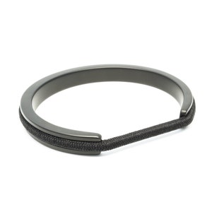 Könnte beinhalten: Ein dunkelgraues Armband mit einem kreisförmigen Metallband und einem schwarzen, strukturierten elastischen Kordel. Das Armband ist ein minimalistisches Design, geeignet für den Alltag. Der Durchmesser beträgt ca. 6,35 cm.