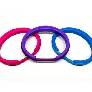 May include: Three colorful hair tie bracelets in pink, purple, and blue are displayed against a white background. The purple bracelet has the word "LOVLO" printed on it. These accessories are designed to be worn on the wrist.