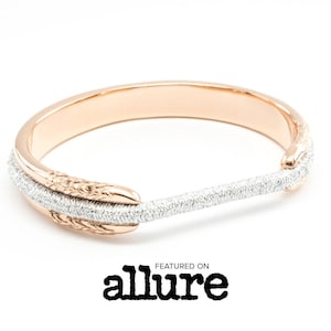 Könnte beinhalten: Roségoldenes und silbernes Armband mit einzigartigem Design. Das Armband hat ein Roségoldband mit strukturierten Details an den Enden und einen silbernen Stab mit kleinen Kristallen. Der Text "FEATURED ON allure" ist darunter.