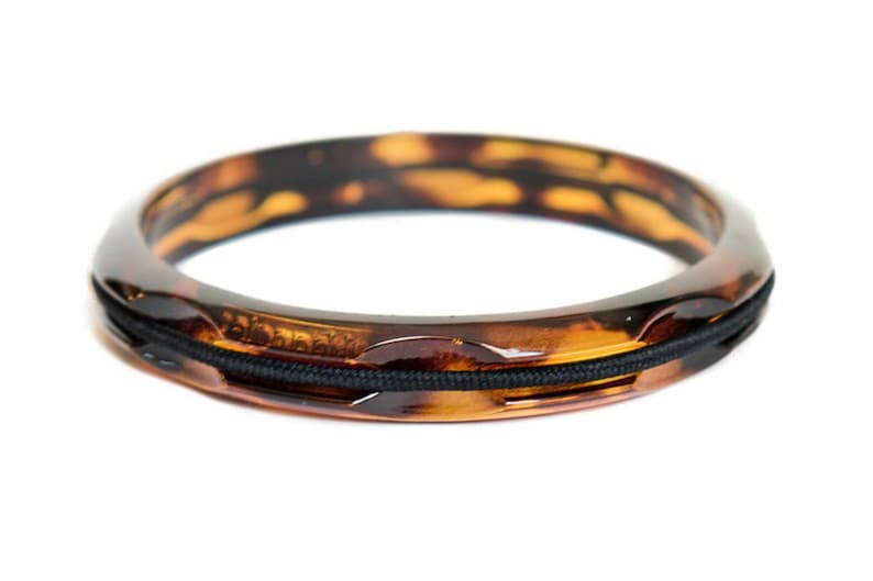 May include: A tortoiseshell-patterned bangle bracelet with a black elastic band detail. The bracelet has a glossy finish and a rounded shape. The warm brown and amber tones create a classic, elegant look.