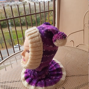 May include: A purple and white knitted hat with a pom-pom on top. The hat has a cowl neck and is made with a chunky yarn.