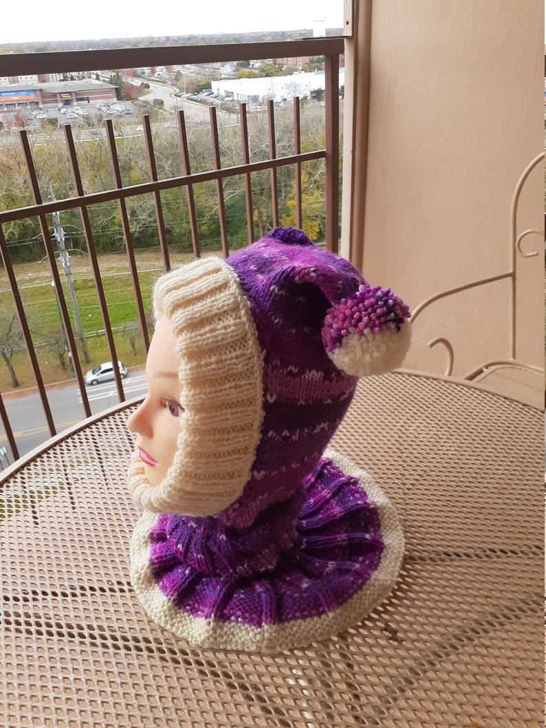 May include: A purple and white knitted hat with a pom-pom on top. The hat has a cowl neck and is made with a chunky yarn.