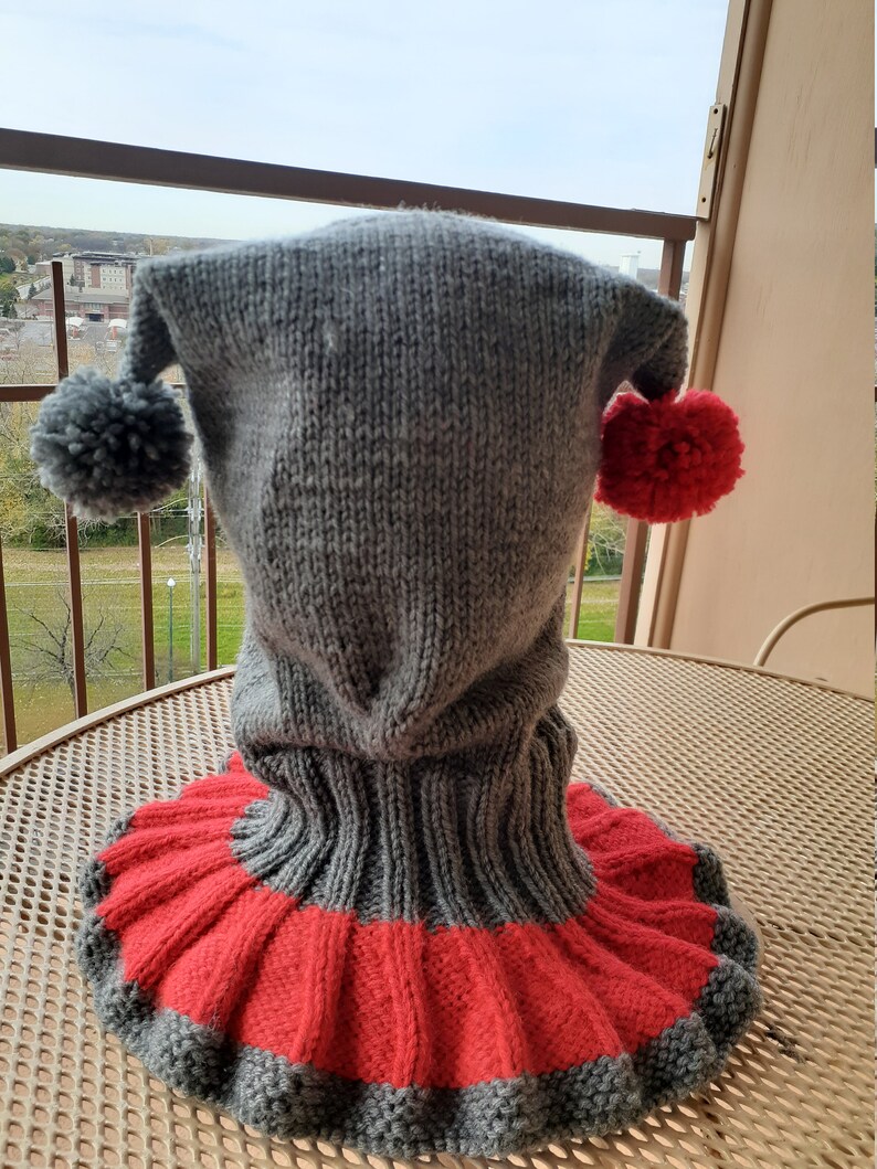Puede incluir: Sombrero de punto gris con un pomp&oacute;n rojo y gris a cada lado. El sombrero tiene un cuello de lana largo que es rojo y gris.