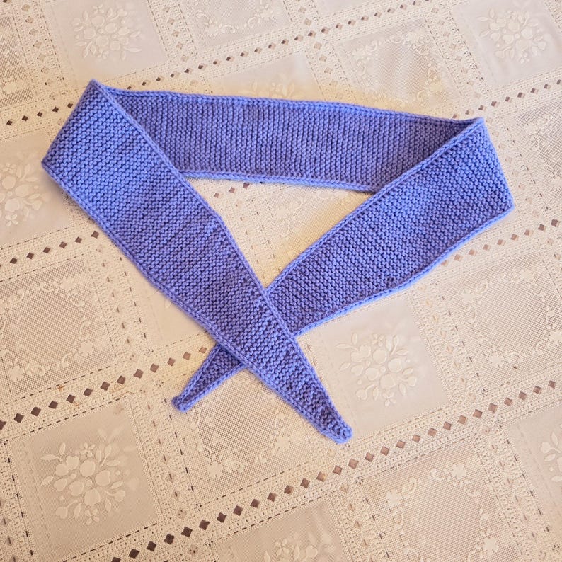 May include: A knitted lavender scarf lies on a white lace tablecloth. The scarf is a solid color and appears to be handmade. The scarf is folded in a V-shape, with the ends pointing downwards. The knitted texture is clearly visible.