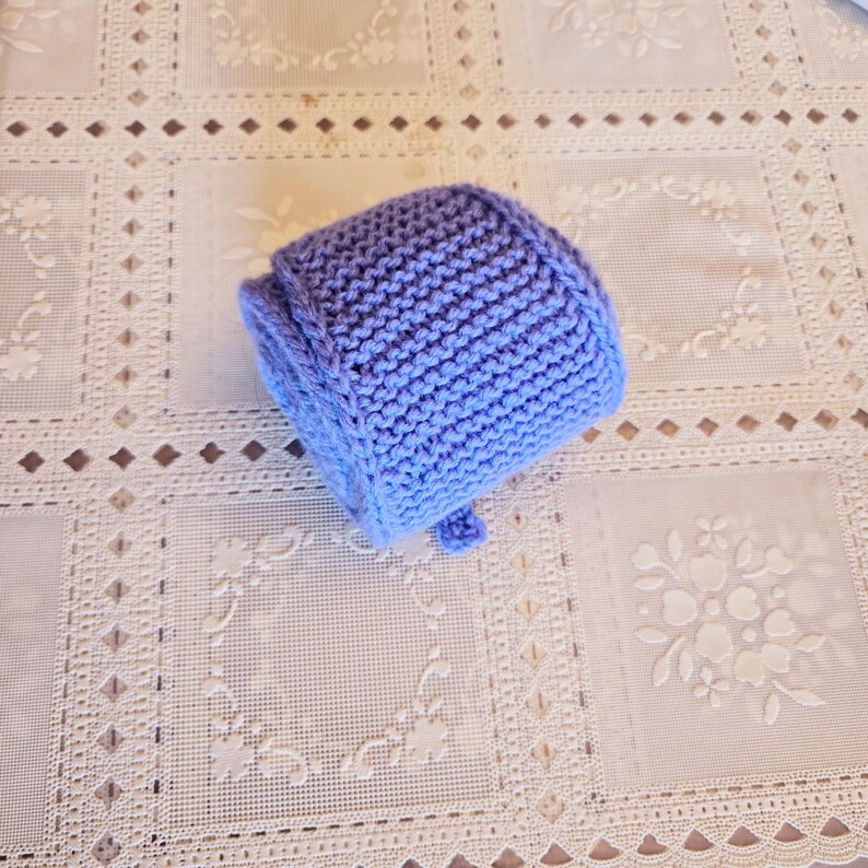 May include: A handmade, knitted baby bonnet in shades of blue. The bonnet features a rounded crown and a small brim, with a textured knit stitch. The bonnet is a soft, pastel blue color, perfect for a newborn.