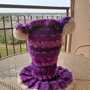 May include: Purple and white knitted hat with pom poms on top. The hat has a long neck warmer attached.
