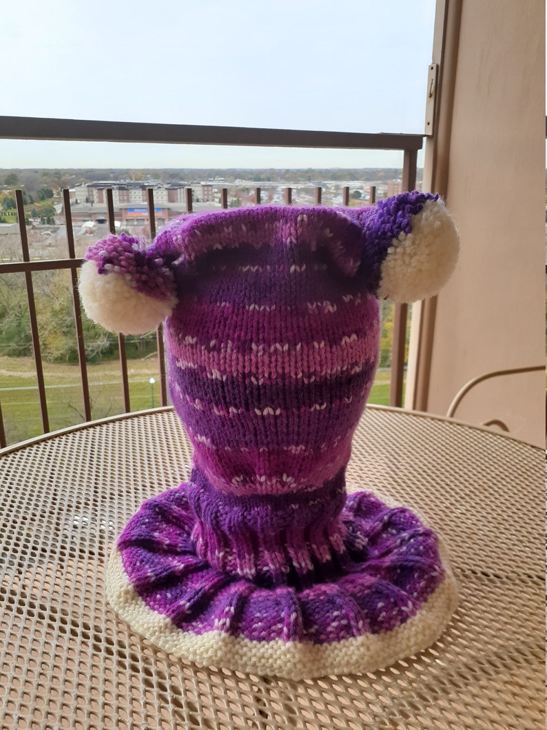 May include: Purple and white knitted hat with pom poms on top. The hat has a long neck warmer attached.