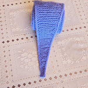 May include: A hand-knitted, light blue scarf with a tapered end. The scarf is made of a soft, textured yarn and is displayed on a cream-colored, patterned tablecloth. The scarf's color is a soft, muted blue.
