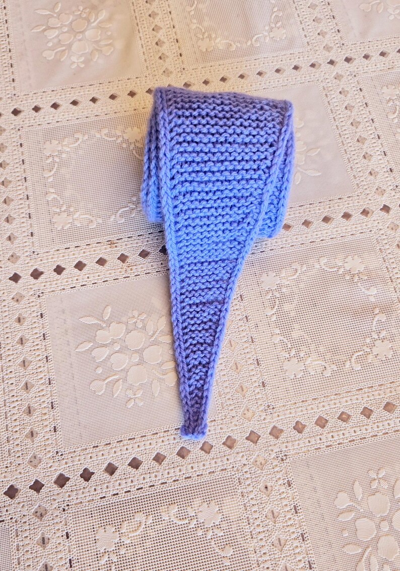 May include: A hand-knitted, light blue scarf with a tapered end. The scarf is made of a soft, textured yarn and is displayed on a cream-colored, patterned tablecloth. The scarf's color is a soft, muted blue.