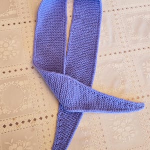 May include: A knitted scarf in a soft periwinkle blue. The scarf is a long, narrow design with a triangular end. The knitted texture is visible, and the scarf appears to be handmade. The scarf is laying on a white patterned surface.