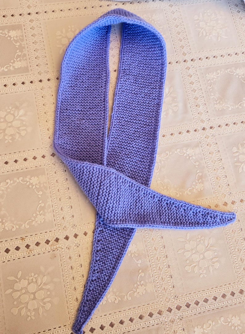May include: A knitted scarf in a soft periwinkle blue. The scarf is a long, narrow design with a triangular end. The knitted texture is visible, and the scarf appears to be handmade. The scarf is laying on a white patterned surface.