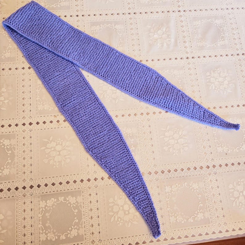May include: A knitted scarf in a soft periwinkle blue hue. The scarf is long and features a pointed end on each side. The knit pattern is visible, adding texture to the accessory. The scarf is laid out on a patterned surface.