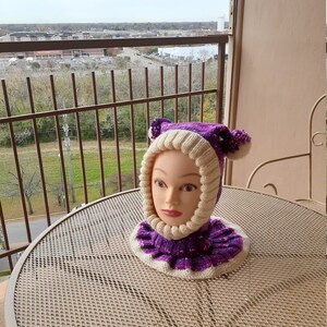 May include: A purple and white knitted hat with ear flaps and a pom-pom on each ear flap. The hat is on a mannequin head.