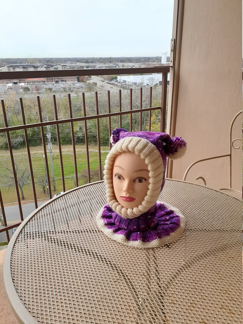 May include: A purple and white knitted hat with ear flaps and a pom-pom on each ear flap. The hat is on a mannequin head.