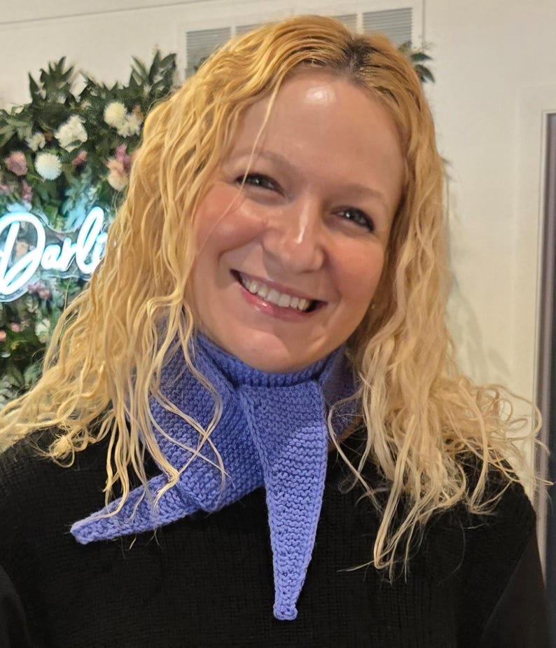 May include: A light purple knitted scarf with a pointed end is worn around the neck. The scarf is made of a soft, textured yarn. The person in the image is wearing a black sweater. The background includes a floral arrangement and a neon sign.