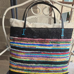 May include: A square tote bag featuring a black crocheted body with horizontal stripes of vibrant multi-colored yarn in shades of blue, yellow, red, and green. The bag has two black leather handles attached with bright blue stitching accents.