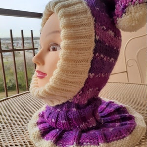 May include: A purple and white knitted hat with a pom-pom on top. The hat has a fold-down brim and a long neck warmer.