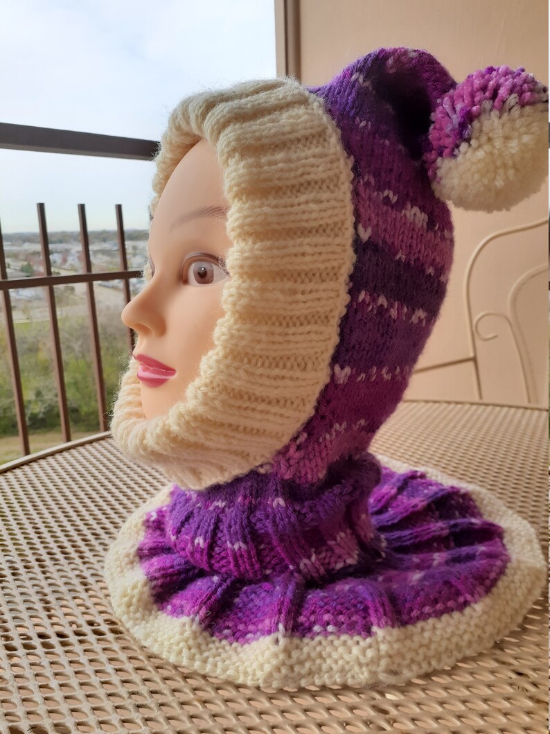 May include: A purple and white knitted hat with a pom-pom on top. The hat has a fold-down brim and a long neck warmer.