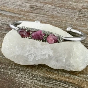 May include: A silver bangle bracelet with a row of pink and gray gemstones set in the metal.